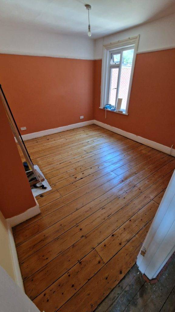 Restored pine floor in a room with orange walls and a large window, showcasing the results of sanding and sealing.